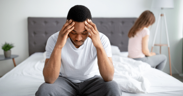 A stressed Black man sits on the edge of a bed holding his head in his hands, while a woman sits turned away from him in the blurred background.