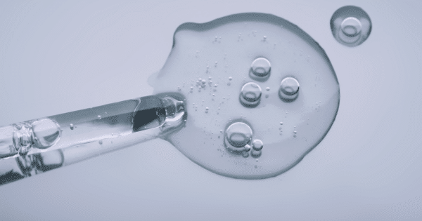 A top-down macro shot of a clear pipette releasing a dollop of transparent gel formula filled with tiny air bubbles onto a clean, light surface.