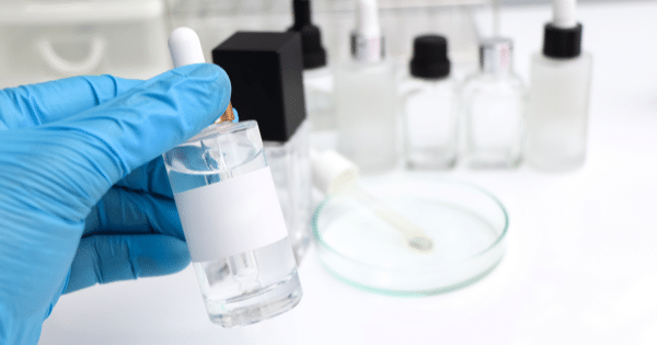 A gloved hand holds up a clear dropper bottle with a blank white label, with a petri dish and several other glass vials blurred in the background.