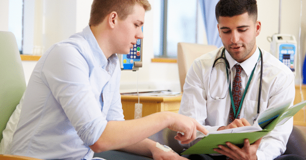 A young male patient sits in a clinic chair and points to a page inside a green medical folder held by a male doctor wearing a tie and a stethoscope.