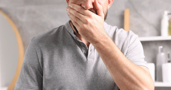 Close-up of a man in a grey polo shirt covering his mouth with his hand, appearing nauseous or about to cough in a bathroom setting.