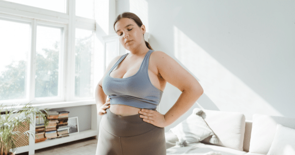 A medium shot of a plus-size woman in a bright, sunlit room, standing with her hands on her hips. She is wearing a blue sports bra and gray leggings, looking downward with a neutral expression.