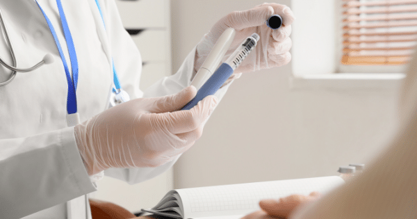 Close-up of a healthcare professional wearing white medical gloves, holding two injection pens—one white and one blue—over a desk with an open notebook.