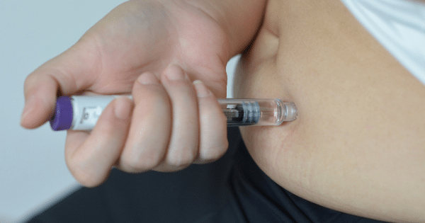 A close-up shot of a person’s hands administering a subcutaneous injection into their abdomen using a clear medicine pen with a purple cap.