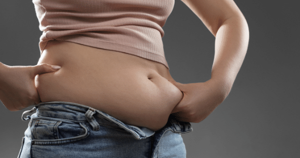 A close-up side view of a person wearing a pink crop top and jeans, using both hands to pinch the skin and fat around their waistline.