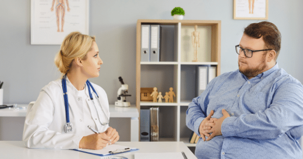 A medical consultation where a female doctor in a white lab coat sitting at a desk listens to a plus-size male patient in a blue shirt who is holding his stomach.