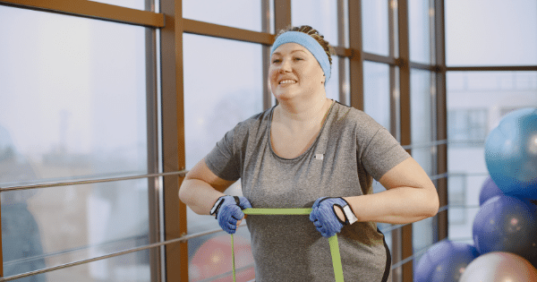 A plus-size woman in a gym setting wearing a gray shirt, blue headband, and workout gloves, smiling while exercising with a green resistance band near large windows.