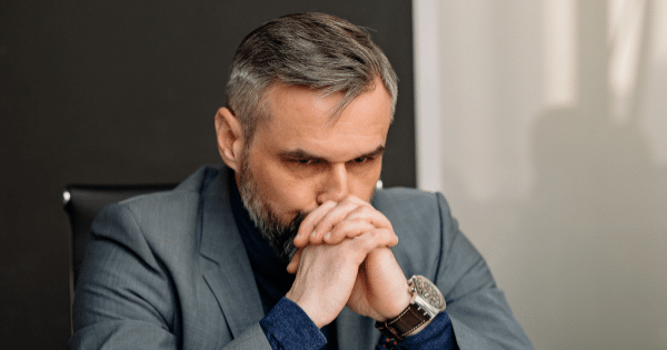 A middle-aged man with graying hair and beard wearing a suit, resting his chin on resting hands in deep thought.