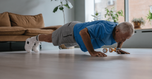 An older man performing a push-up exercise on a wooden living room floor next to small dumbbells.