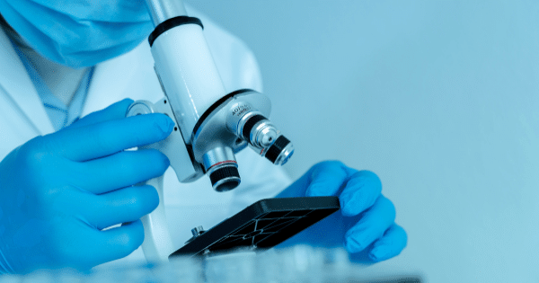 Close-up of a scientist wearing blue rubber gloves, a face mask, and a lab coat adjusting a white light microscope.