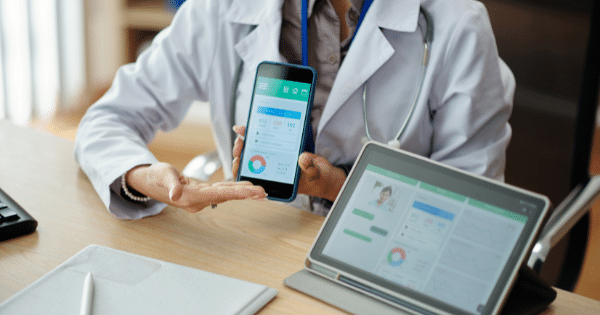 A doctor sitting at a desk presenting a smartphone screen displaying health data charts, alongside a tablet showing similar medical analytics.