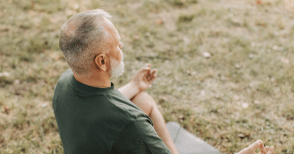 High-angle view of a senior man with gray hair and beard meditating outdoors in a cross-legged lotus posture on a grassy lawn.
