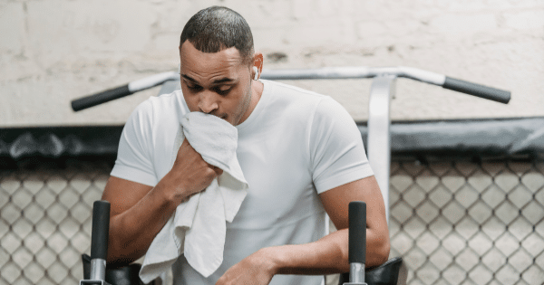 Man in a white athletic shirt resting his chin against a white towel to wipe sweat during a gym workout break.