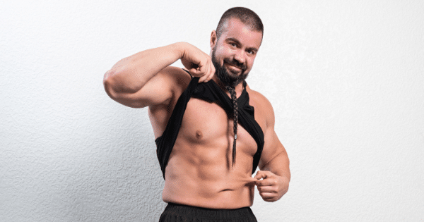 Muscular man with a braided beard lifting his black tank top to show off his abdominal muscles while pinching a bit of skin at his side.