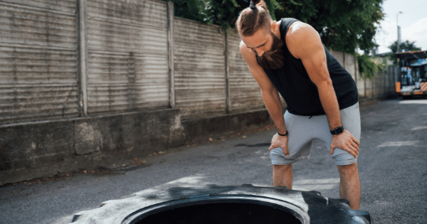 Athletic man with a long beard and topknot hairstyle leaning forward with hands on his knees, catching his breath next to a heavy workout tire outdoors.