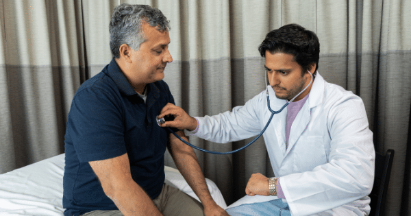 A young male doctor in a white lab coat using a stethoscope to check the heartbeat of a middle-aged male patient sitting on an examination table.