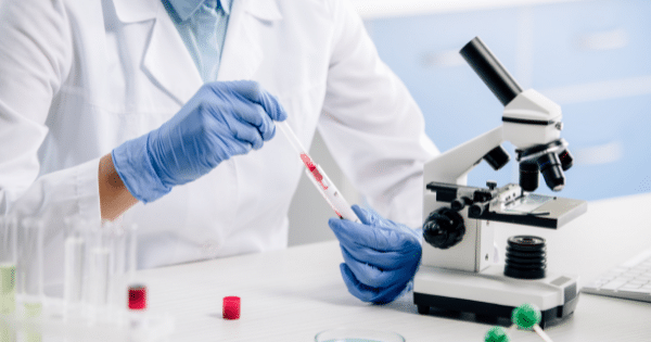 A laboratory technician in a white coat and blue surgical gloves holding a blood sample test tube next to a optical microscope.