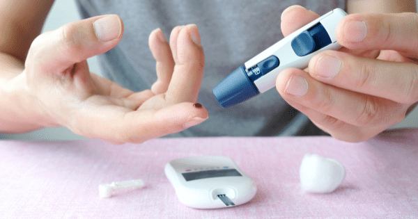 A person using a blue lancing device to prick their finger for a blood sugar glucose test, with a blood drop visible alongside a digital monitor.