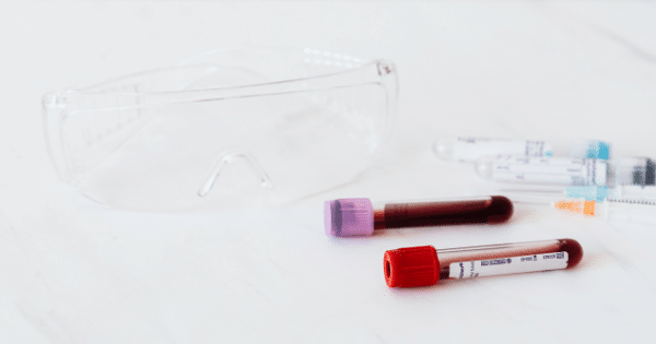 Protective laboratory safety glasses resting next to two blood sample test tubes and medical syringes on a clean white surface.