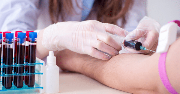 A healthcare worker wearing white gloves drawing blood from a patient's arm using a syringe, next to a rack filled with blood test tubes.