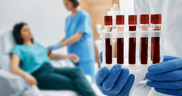 Gloved hands holding a rack of blood sample vials in the foreground, with a healthcare worker drawing blood from a patient blurred in the background.