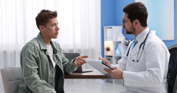 A young man sitting in a clinic consultation room speaking gesturingly to a doctor wearing a white lab coat and stethoscope who holds a clipboard.