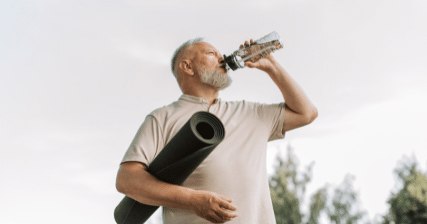 A low-angle shot of a middle-aged man with a grey beard drinking water from a clear bottle while holding a rolled-up black exercise mat under his arm outdoors.