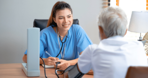 A smiling female healthcare provider in blue scrubs taking an older man's blood pressure using a sphygmomanometer across a clinic desk.