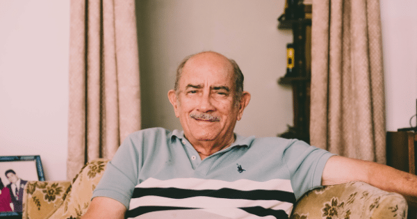 An older man with a mustache wearing a striped polo shirt sitting comfortably on a sofa and smiling at the camera.
