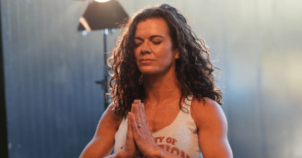 A fit woman with long curly dark hair holding her hands together in a prayer pose with closed eyes in a softly lit gym.