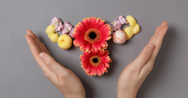 Two open hands framing a flat-lay arrangement of colorful flowers laid out in the shape of a female reproductive system on a grey background.