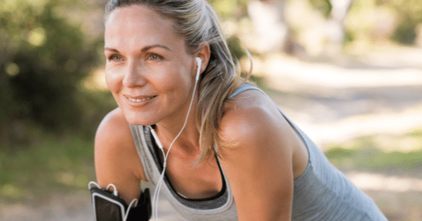 A smiling athletic woman with blonde hair in a ponytail wearing earphones and leaning forward outdoors.