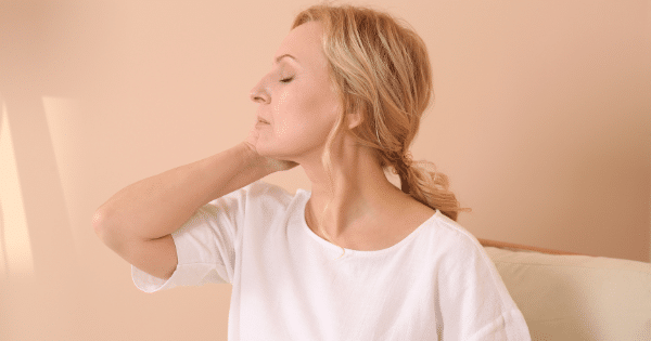 A middle-aged blonde woman sitting on a couch with her eyes closed while gently rubbing her neck.