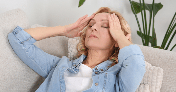 A blonde woman in a denim shirt lying back on a couch with eyes closed, pressing her hands against her temples in pain or stress.