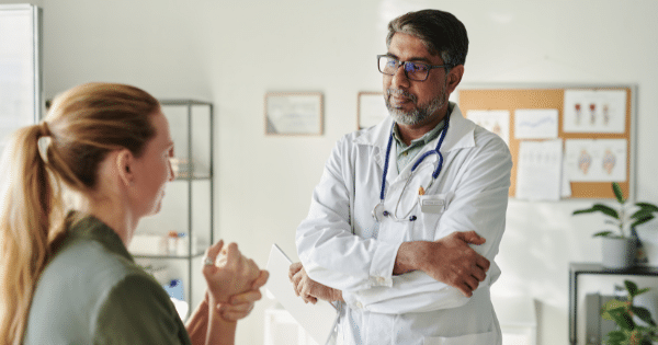 A male doctor with glasses and a stethoscope standing with crossed arms listening to a patient in an office.