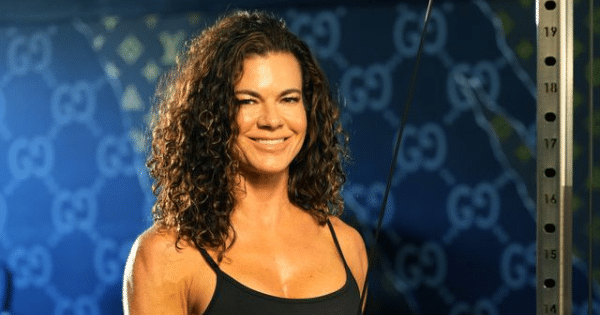 A smiling woman with curly hair standing next to a cable pulley gym machine against a branded blue backdrop.