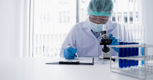 Laboratory scientist in protective gear taking notes near a microscope and test tubes filled with blue liquid.