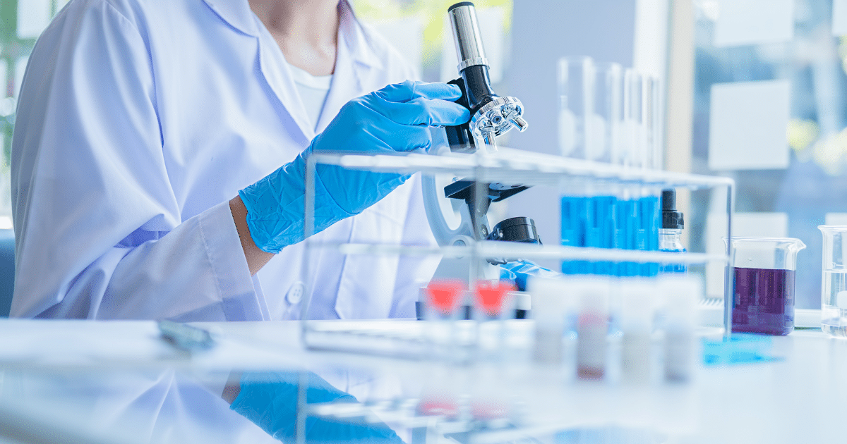 A scientist in a white lab coat and blue protective gloves adjusts a microscope at a laboratory workstation surrounded by test tubes filled with blue liquid and vials.