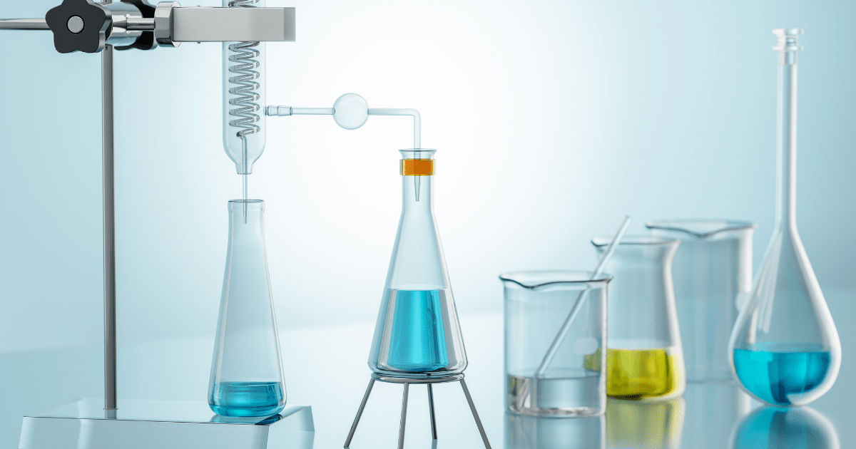 A clean chemical laboratory setup featuring clear glass labware filled with blue and yellow liquids, including Erlenmeyer flasks, a condenser tube, a glass beaker with a stirring rod, and a volumetric flask on a light blue background.