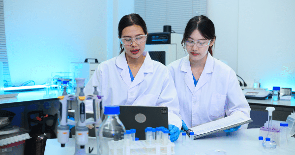 Two female lab researchers wearing lab coats and safety glasses examining data on a tablet.
