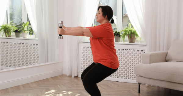 A woman in workout attire performing squat exercises with dumbbells in a living room.