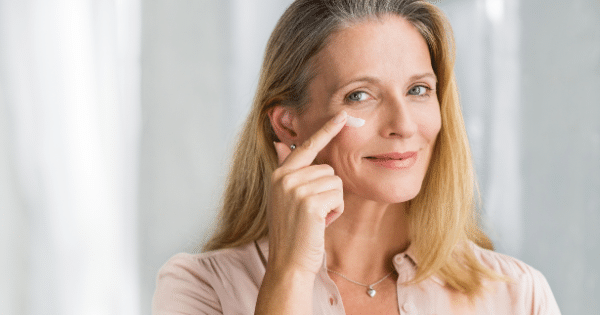 A middle-aged woman with blonde and grey hair smiling gently while using her index finger to apply a dab of white skincare cream under her right eye.