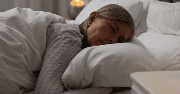 A middle-aged woman with long blonde hair sleeping soundly on her stomach in bed, her head resting comfortably on a fluffy white pillow under a white duvet.