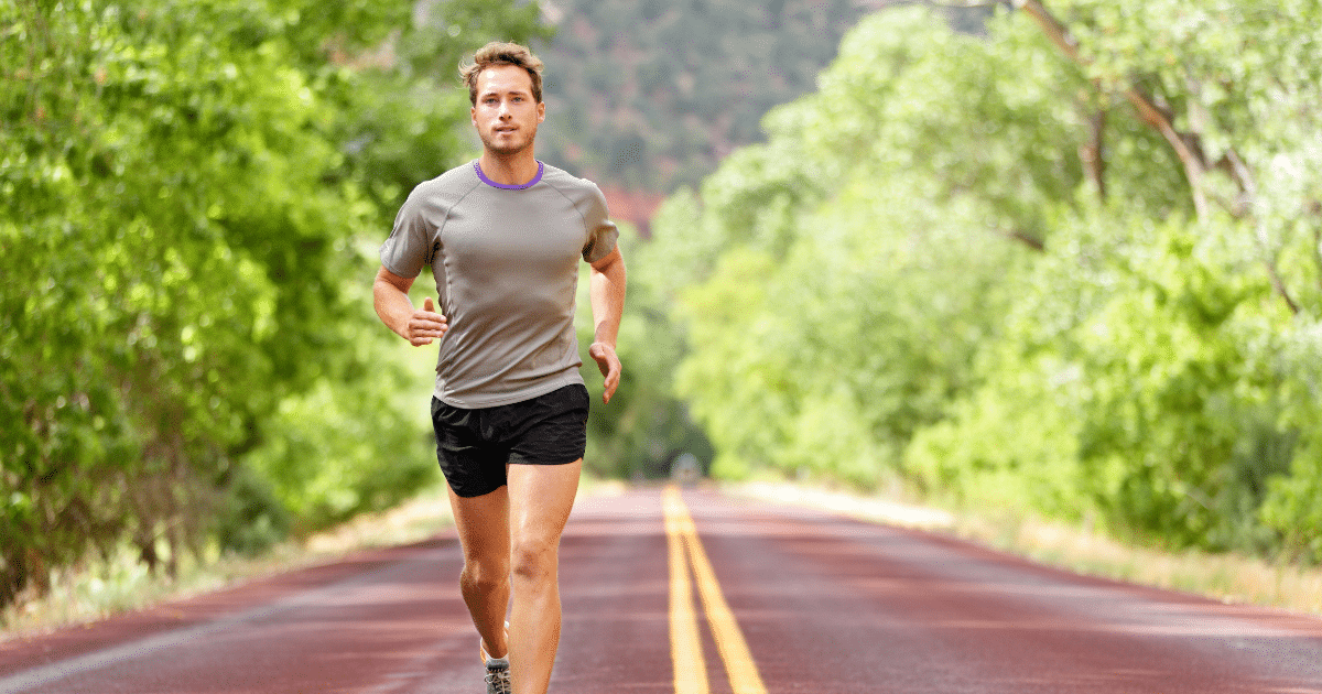 A young athletic man running toward the camera down the center of an outdoor paved road lined with green trees.
