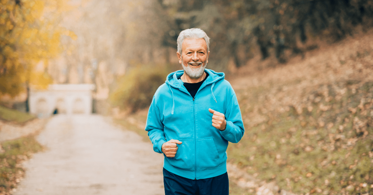 A smiling senior man with white hair and a beard wearing a light blue hooded sweatshirt while jogging along a park path.