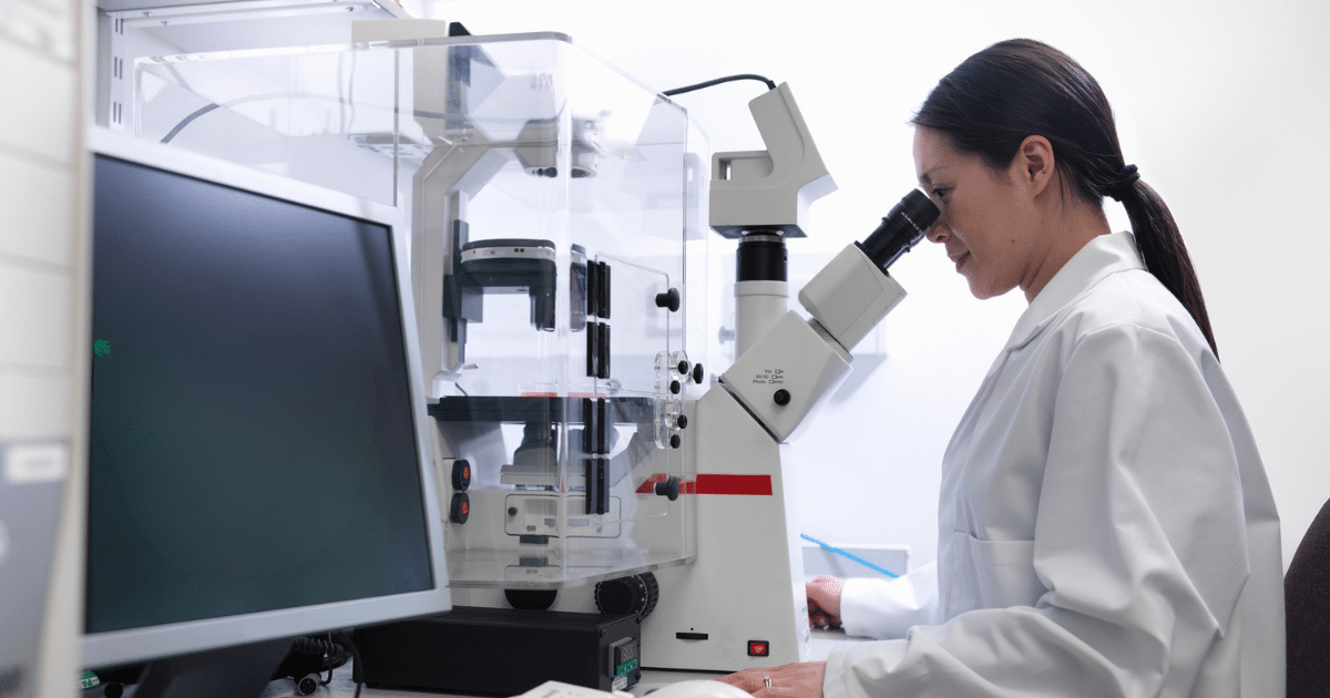 Side profile of a female scientist wearing a white lab coat with her hair tied in a ponytail, looking into the eyepiece of a large, complex laboratory microscope in a bright clinical setting.