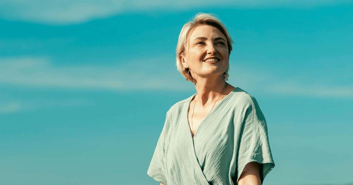 A blonde woman in a sage green wrap dress smiling warmly while looking off-camera against a clear blue sky.