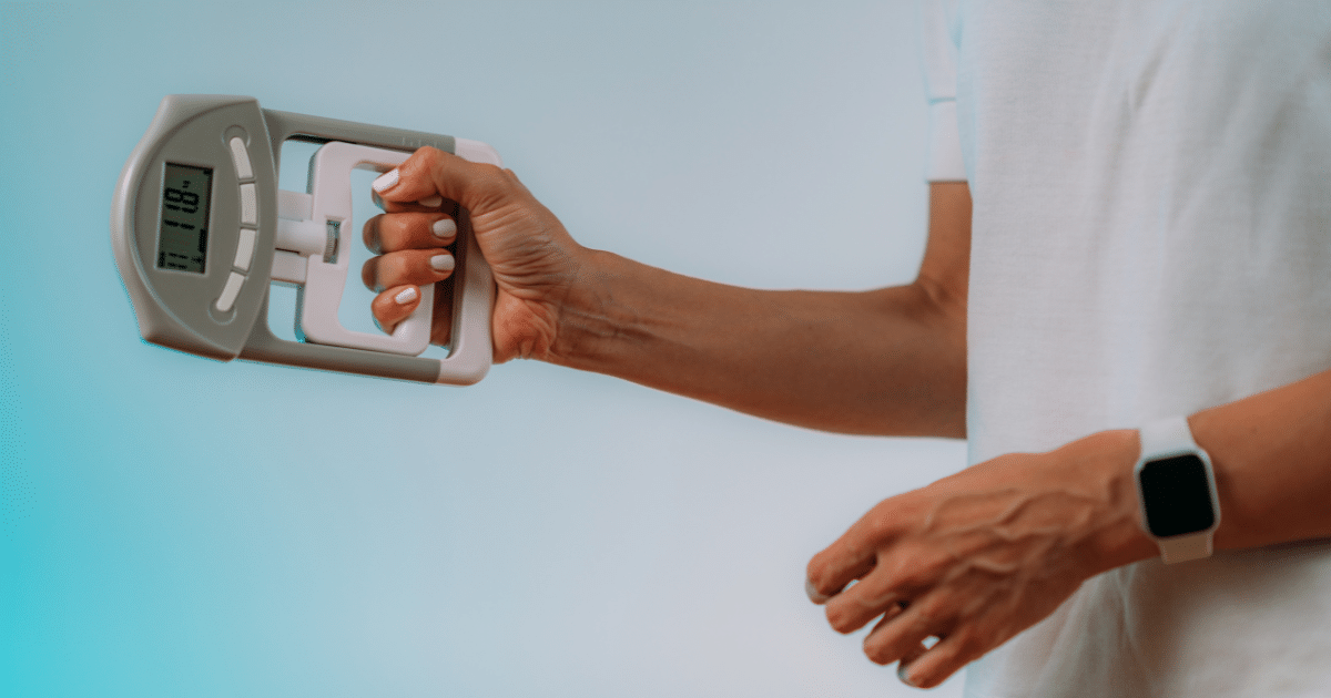 Side view of a person wearing a white t-shirt squeezing a gray digital hand dynamometer with a small LCD screen against a dual-tone blue and white background.