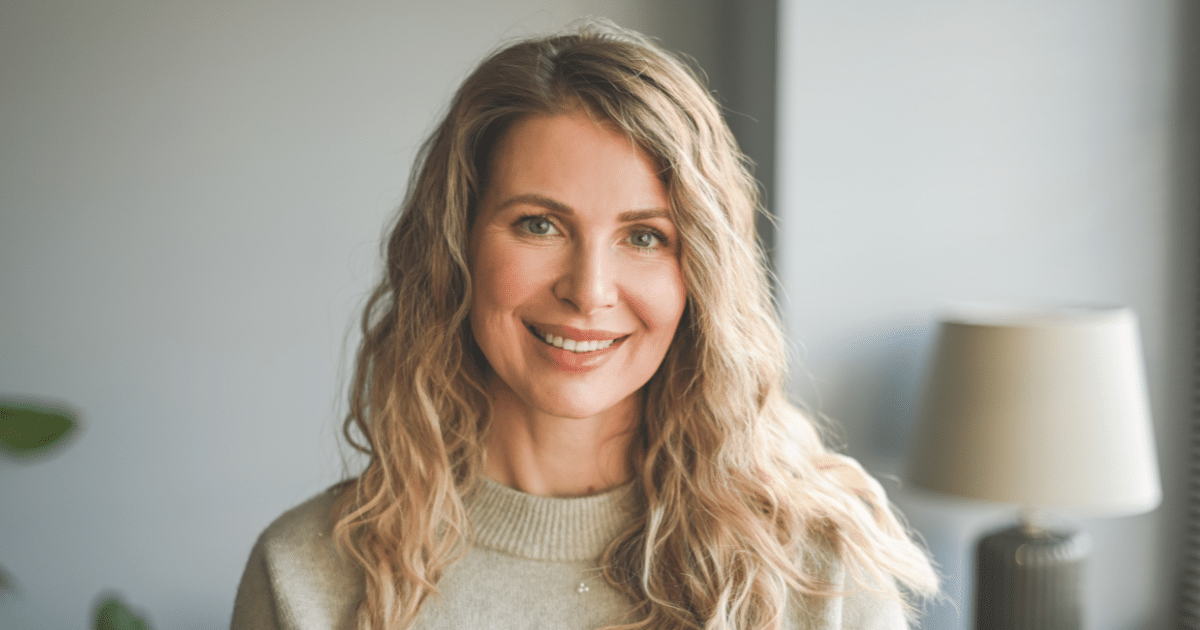 A close-up portrait of a middle-aged woman with wavy blonde hair, smiling softly at the camera. She is wearing a light green knit sweater inside a brightly lit room with a lamp blurred in the background.