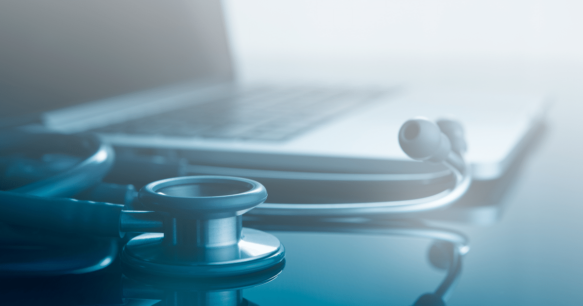A close-up, shallow depth-of-field shot of a silver stethoscope sitting on a dark, reflective surface in the foreground, with a blurred laptop open in the background under bright, clean lighting.